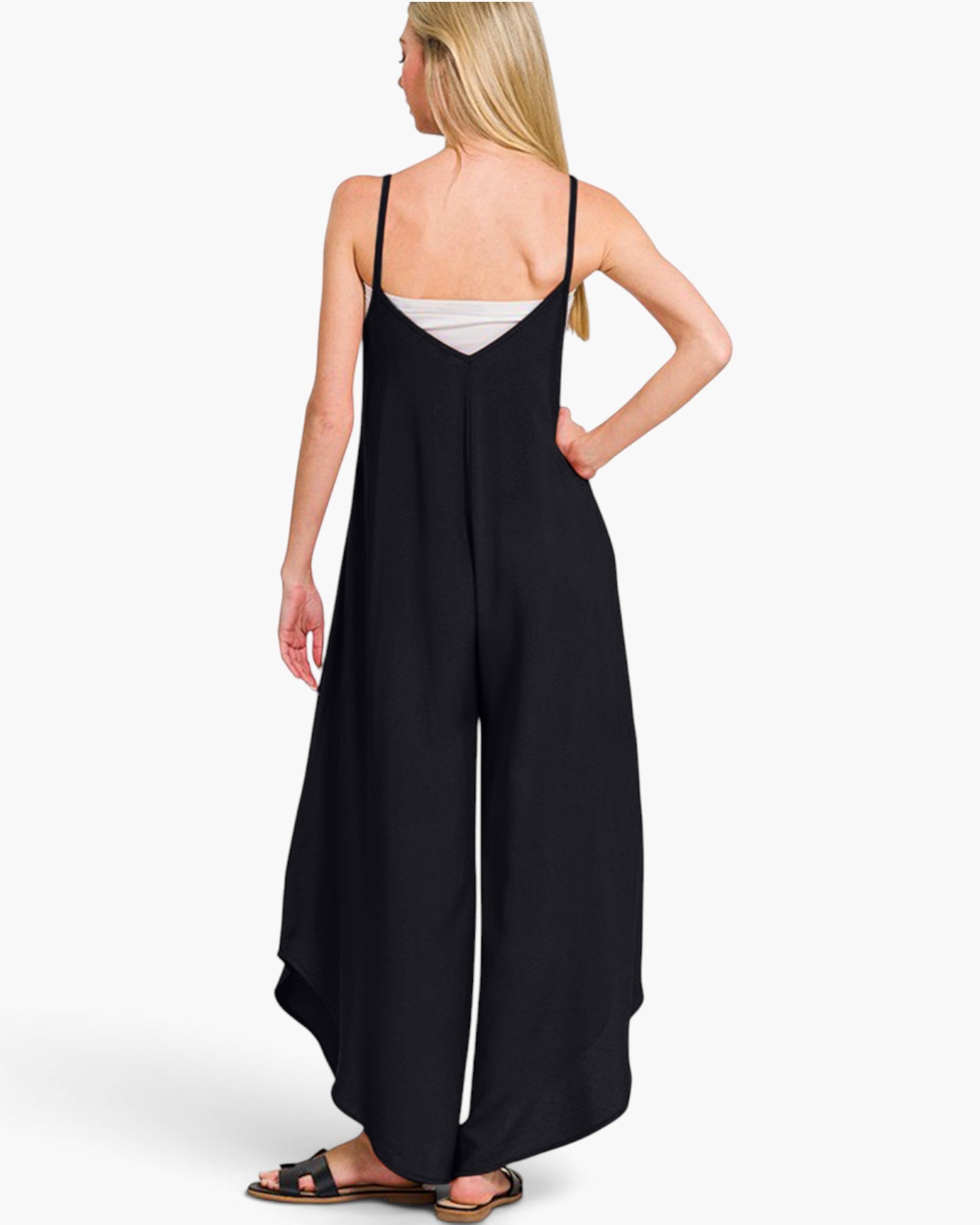 A woman shown from the back wearing a black wide-leg jumpsuit with thin spaghetti straps over a white camisole, paired with black sandals, against a plain white studio background.