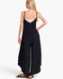A woman shown from the back wearing a black wide-leg jumpsuit with thin spaghetti straps over a white camisole, paired with black sandals, against a plain white studio background.