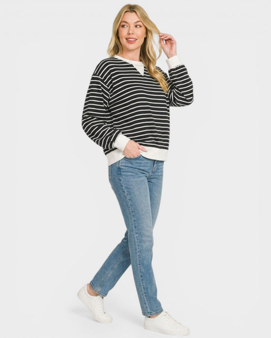 Woman wearing a black and white striped sweater with a white shirt underneath, blue jeans, and white sneakers on a light gray background.