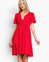 A woman modeling a bright red short-sleeve wrap-style dress with a V-neckline and gathered waist, photographed against a plain light gray studio background.