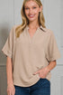 Woman wearing a beige short-sleeve shirt with a collar against a gray wall.