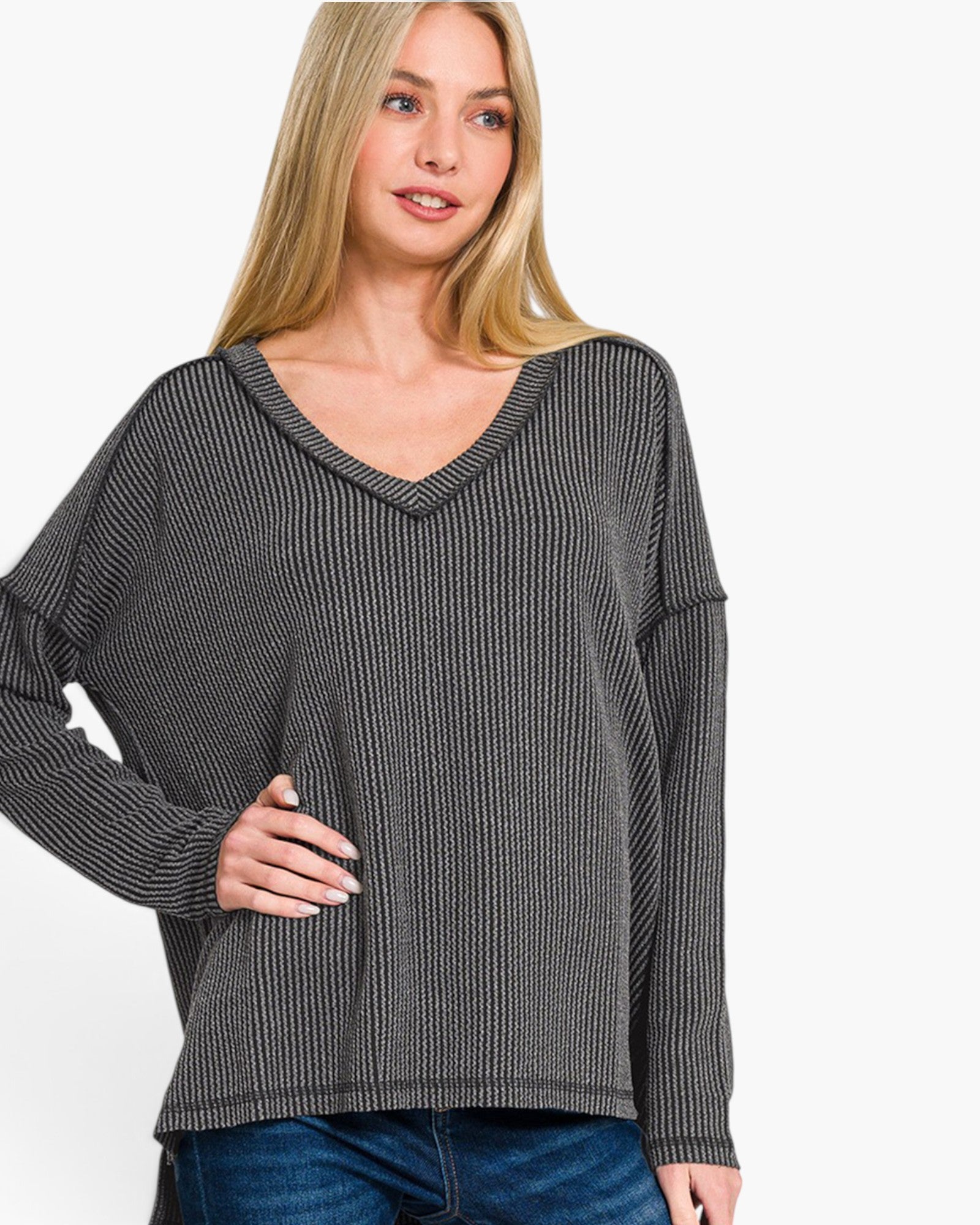 A woman wearing a dark gray ribbed knit long-sleeve sweater with a V-neck, paired with blue jeans, against a plain white background.