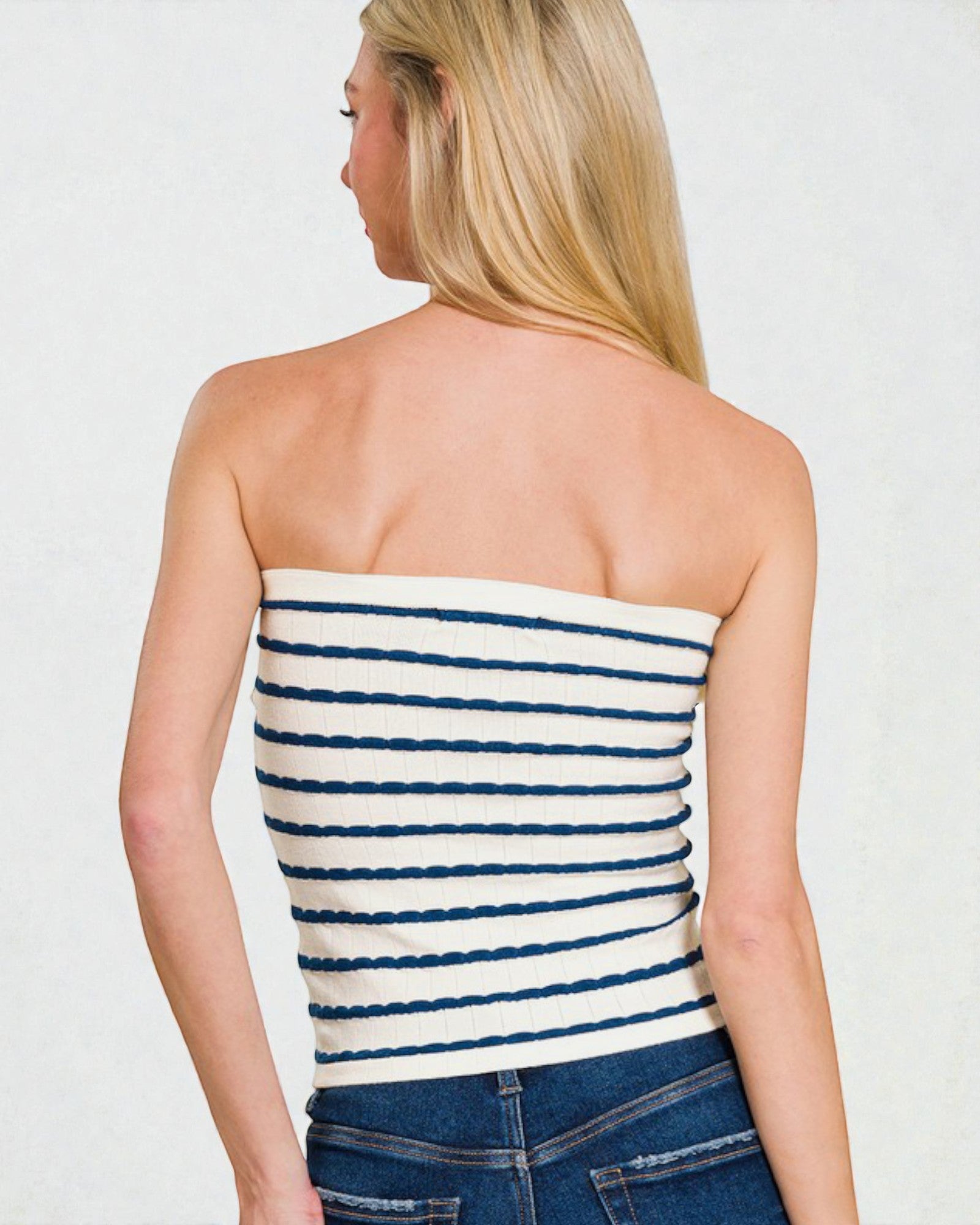 Woman wearing a strapless navy and white striped top with blue jeans on a plain background