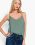 A woman wearing a sage green spaghetti-strap camisole top and dark blue jeans, posing against a clean white studio background.