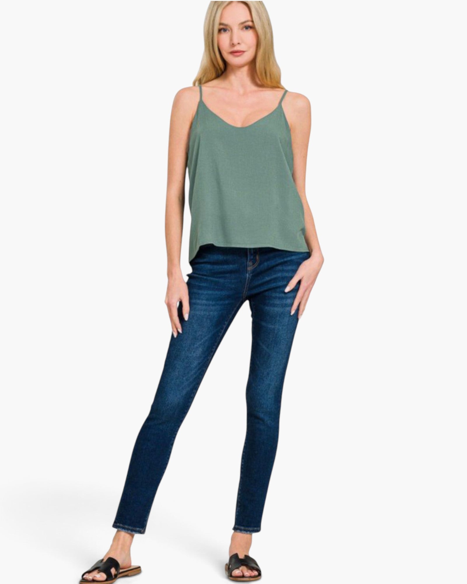 A woman modeling a sage green spaghetti-strap camisole top paired with dark blue skinny jeans and brown slide sandals, set against a plain white studio background.