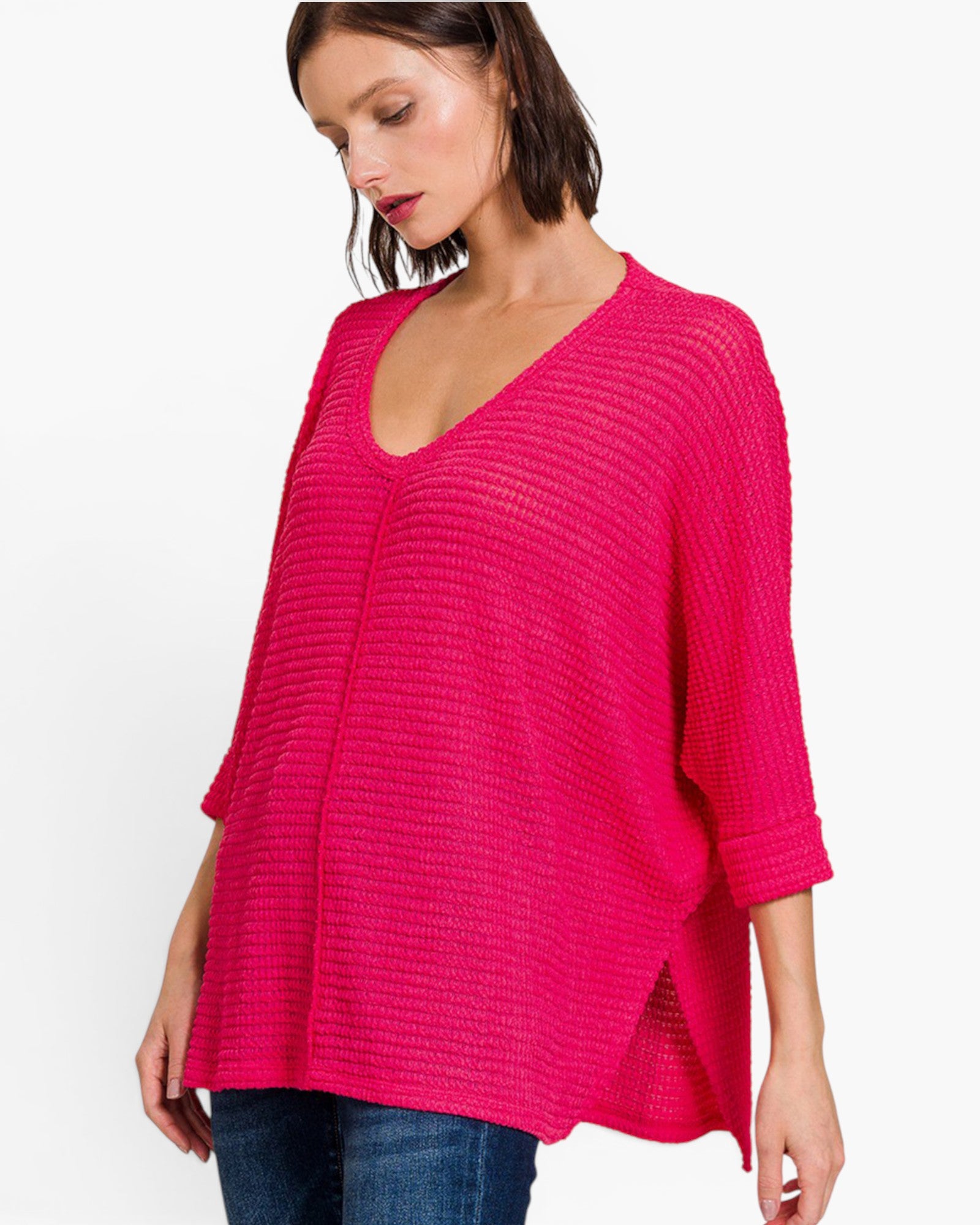 A woman wearing a bright pink ribbed knit top with a V-neck and elbow-length sleeves, paired with blue jeans, against a plain white background.