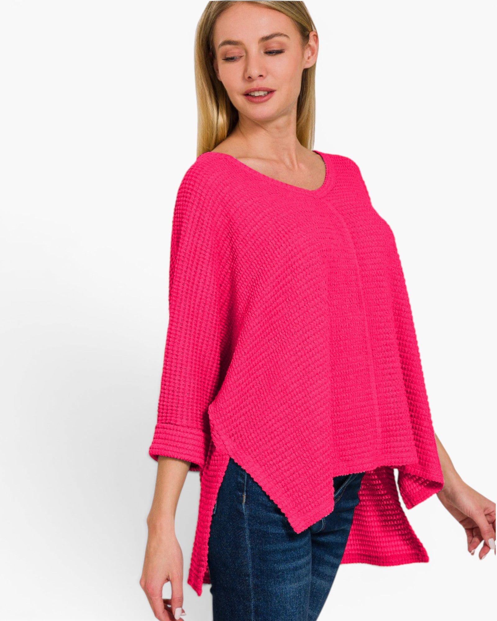 A woman wearing a bright pink waffle-knit poncho top with three-quarter sleeves and an asymmetrical hem, paired with dark blue jeans, against a plain white studio background.