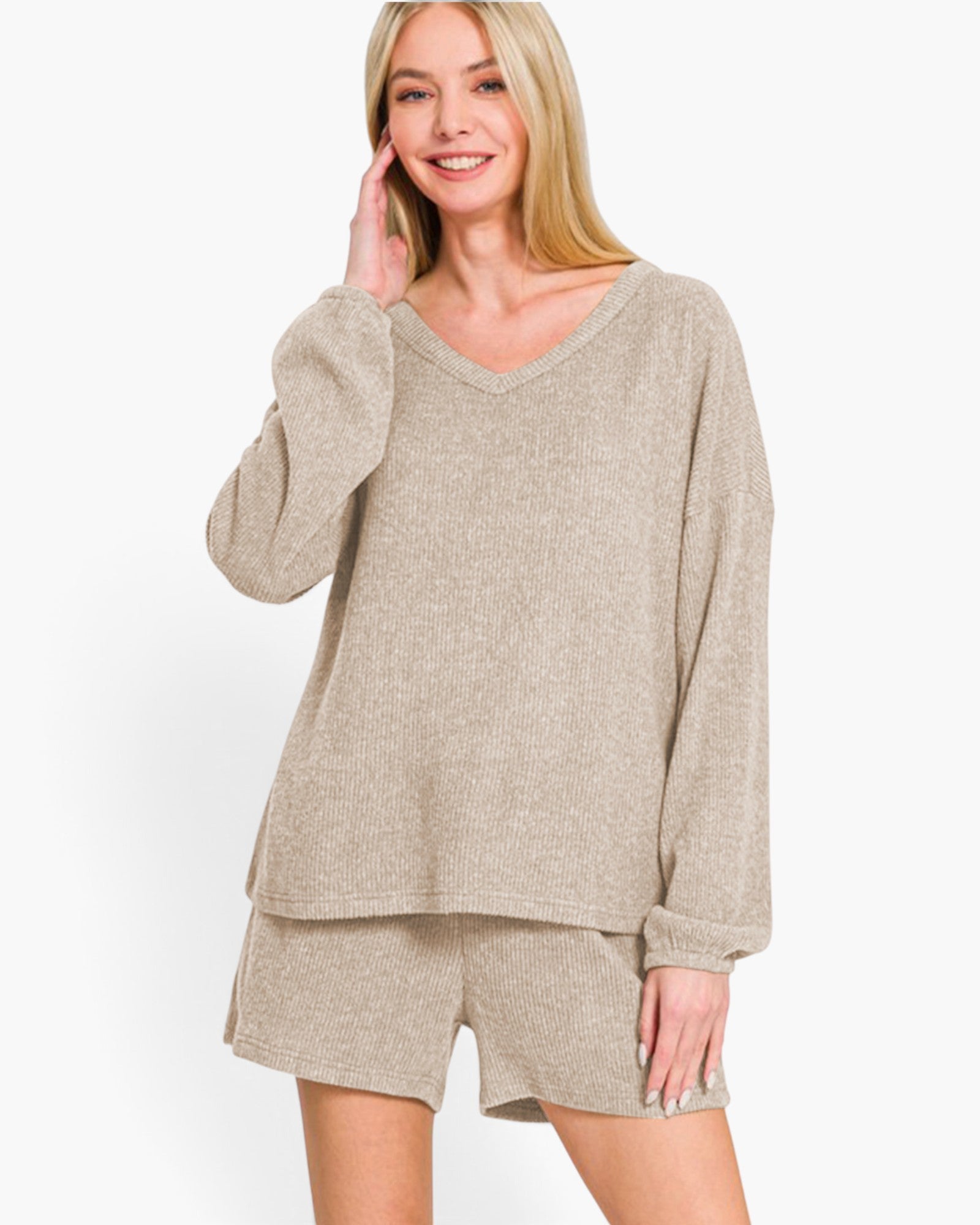 A woman modeling a matching beige lounge set consisting of a long-sleeve V-neck knit top and coordinating shorts, photographed against a clean white studio background.