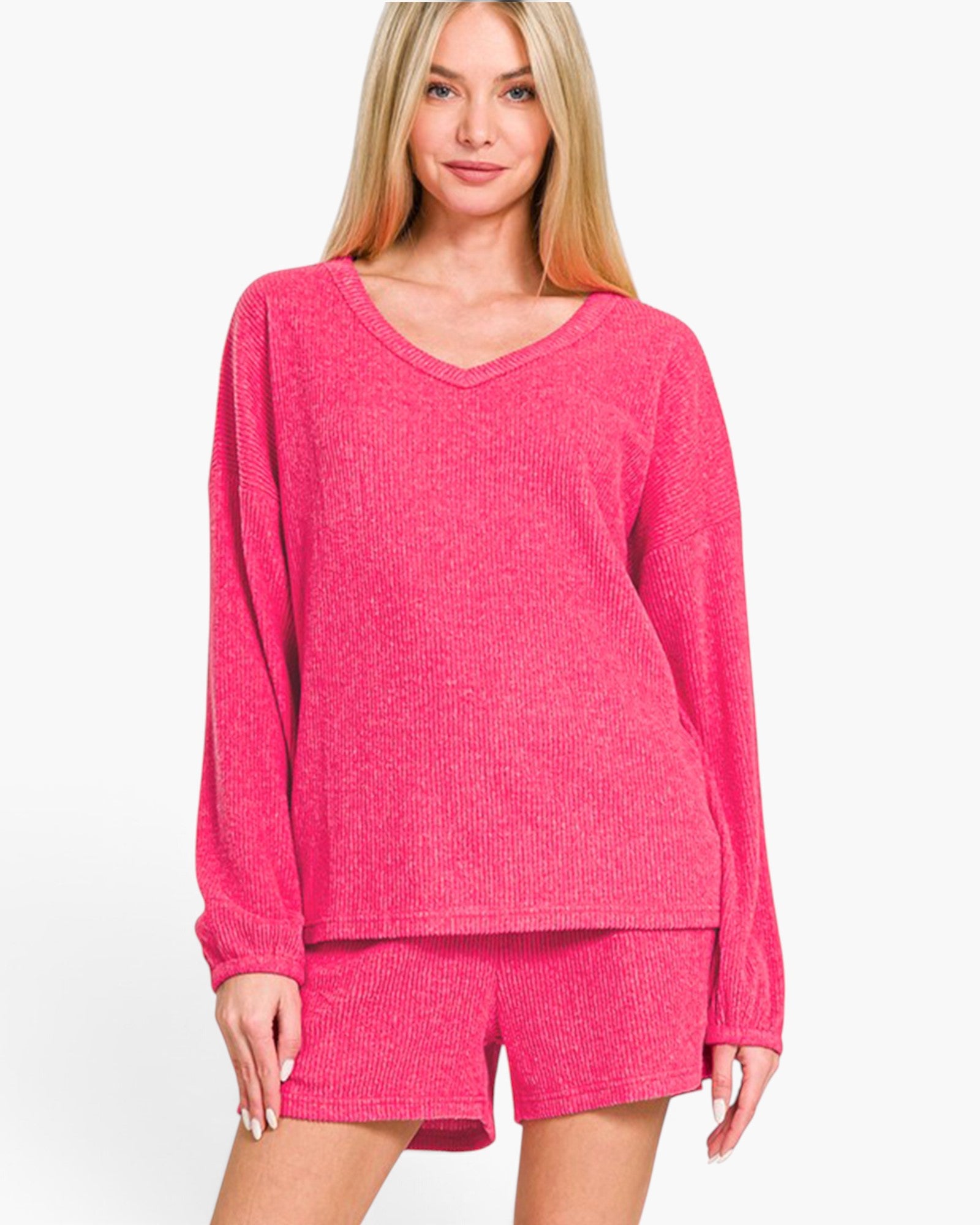 A woman modeling a bright pink knit lounge set consisting of a long-sleeve V-neck top and matching shorts against a plain white background.