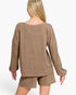 A woman shown from the back wearing a loose, heathered taupe-brown knit sweater and matching shorts against a plain light gray studio background.