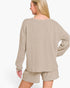 A woman shown from the back wearing a beige ribbed knit lounge set consisting of a long-sleeve top and matching shorts against a plain white background.