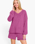 A woman modeling a matching two-piece loungewear set in a heathered magenta-purple color, featuring a long-sleeve V-neck top and coordinating shorts, against a plain white studio background.