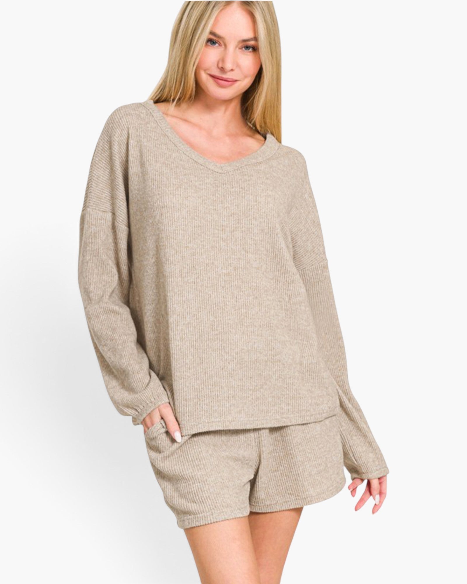 A woman modeling a matching knit loungewear set consisting of a long-sleeve v-neck sweater and coordinating shorts in a heathered light beige color, set against a plain white studio background.