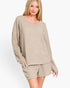 A woman modeling a matching knit loungewear set consisting of a long-sleeve v-neck sweater and coordinating shorts in a heathered light beige color, set against a plain white studio background.