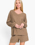A woman modeling a matching lounge set consisting of a long-sleeve v-neck knit top and coordinating shorts in a heathered taupe-brown color, set against a plain white studio background.