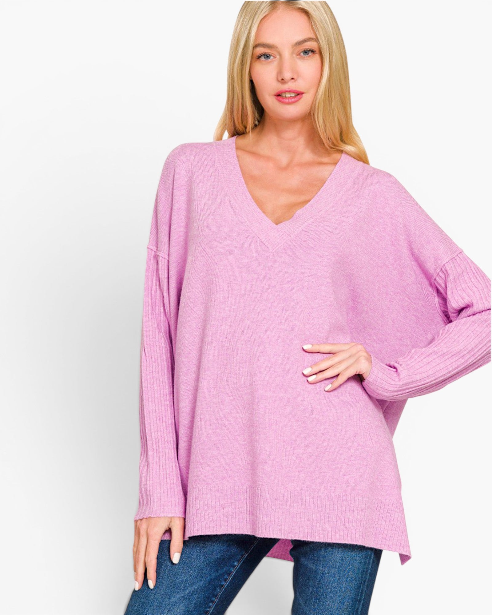 A woman wearing an oversized light pink knit sweater with a V-neck and dropped shoulders, paired with blue jeans, against a plain white studio background.
