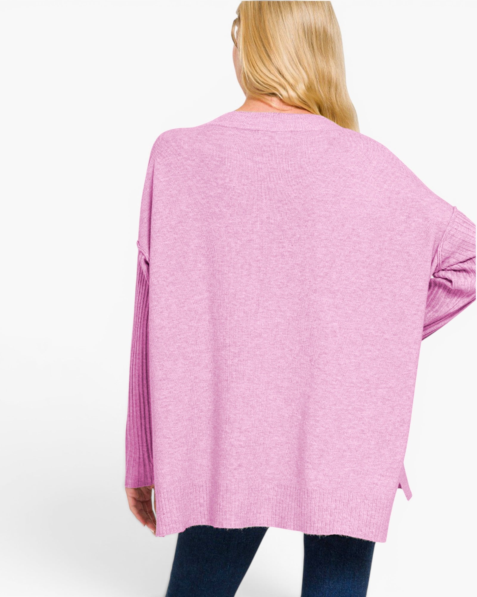 A woman shown from the back wearing a loose, oversized pink knit sweater with ribbed sleeves and side slits, paired with dark blue jeans against a plain white studio background.