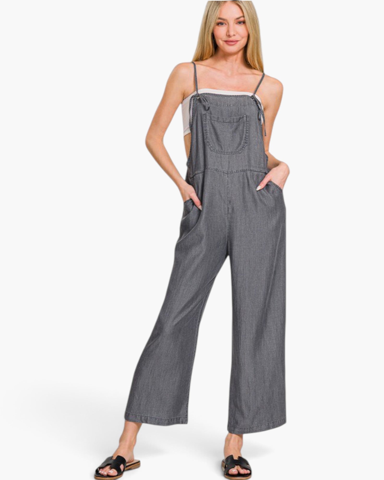 A woman modeling a gray wide-leg jumpsuit with thin spaghetti straps, front pockets, and a button-up bib, styled over a white camisole and black slide sandals against a plain white background.