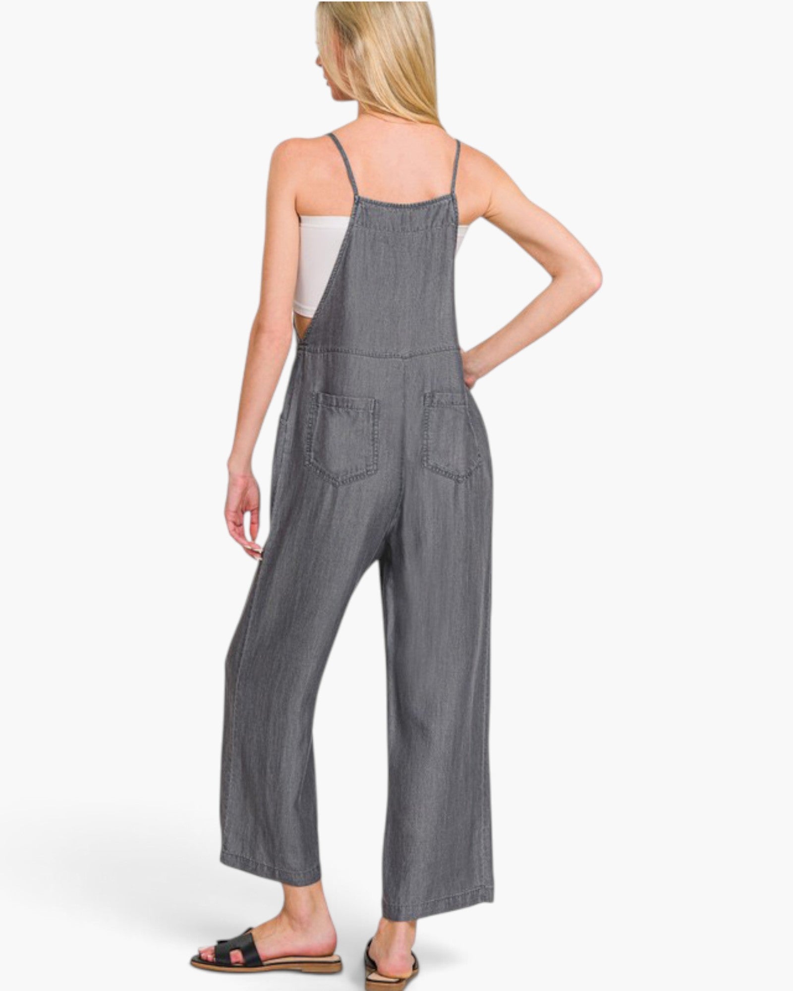 A woman shown from the back wearing a gray wide-leg denim jumpsuit over a white camisole, paired with black slide sandals, against a plain white studio background.