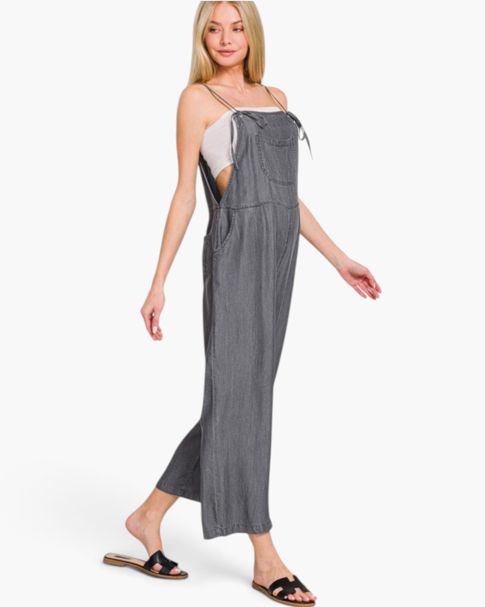 A woman modeling a gray sleeveless wide-leg jumpsuit with thin straps and side cutouts, worn over a white camisole and black slide sandals, against a plain white background.