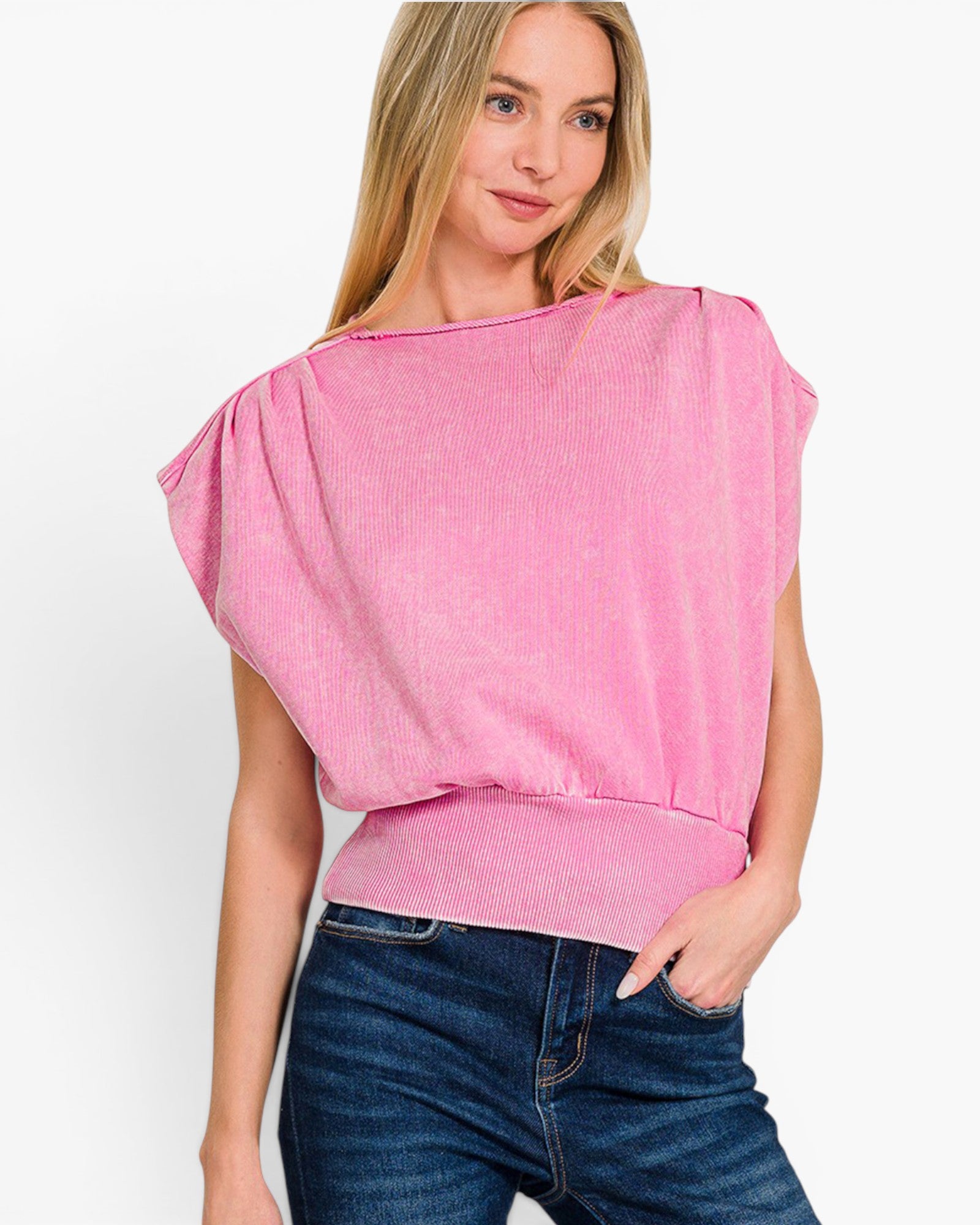 A woman wearing a pink textured knit top with a wide boat neckline and draped cap sleeves, paired with dark blue jeans, against a plain white background.