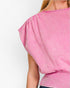 A close-up of a person wearing a pink ribbed knit top with a wide boat neckline and voluminous draped short sleeves, shown against a plain light background.