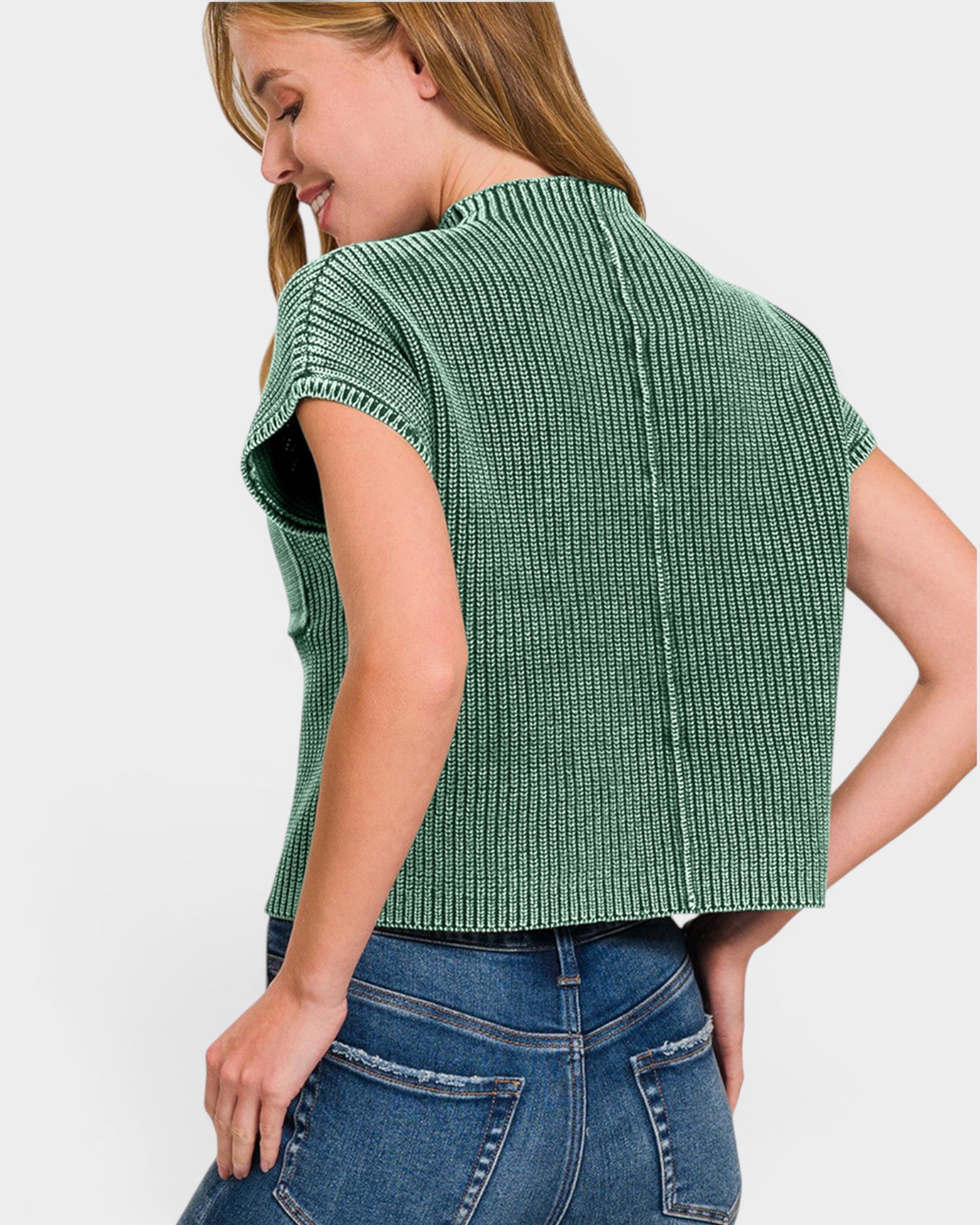 A woman wearing a green ribbed short-sleeve crop top and blue jeans, shown from a back-three-quarter angle against a light gray studio background.