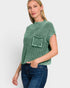 A woman wearing a green ribbed knit short-sleeve top with a chest pocket, paired with dark blue jeans, against a light gray studio background.