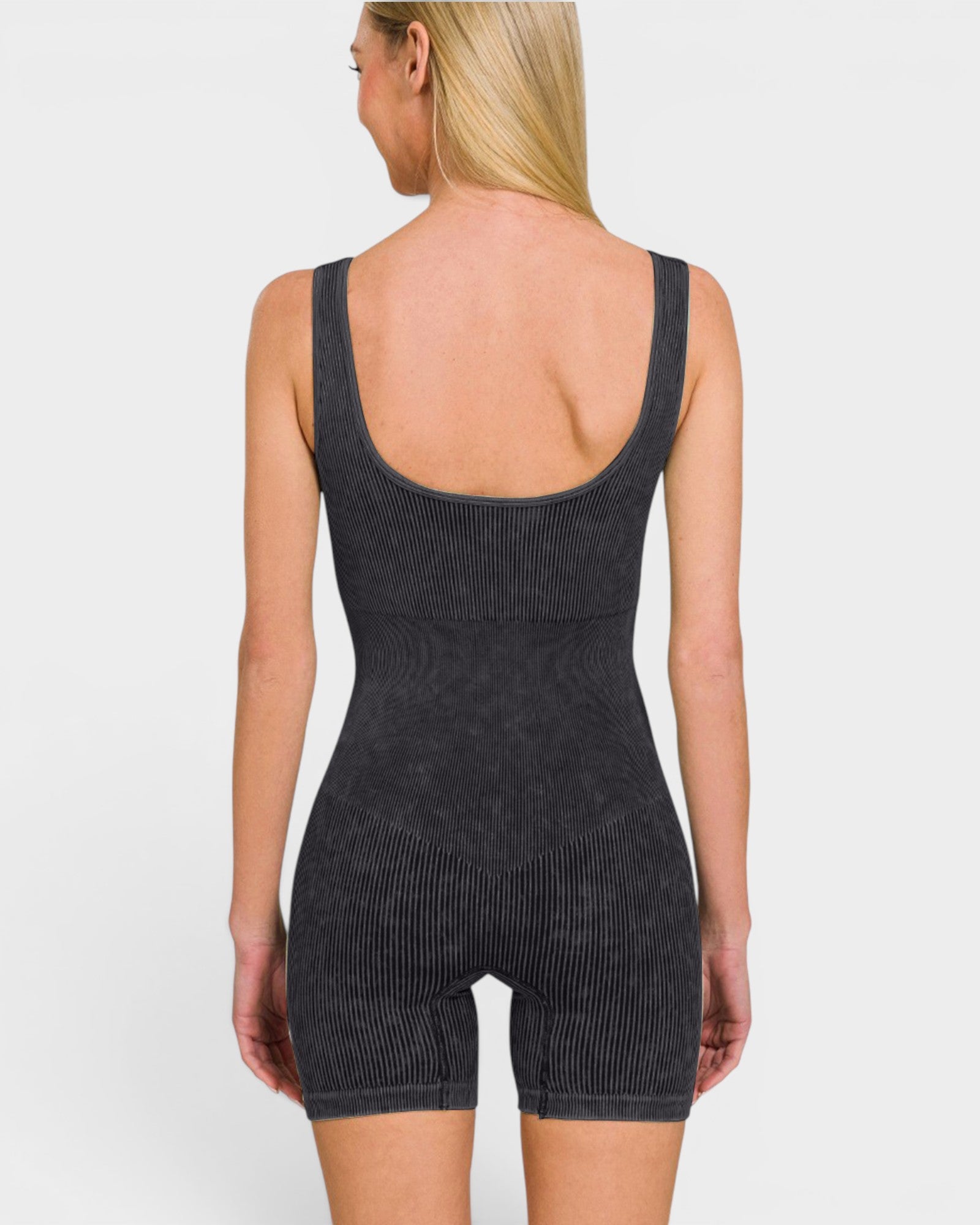 A woman shown from the back wearing a dark charcoal gray ribbed one-piece romper with shoulder straps and mid-thigh shorts, against a light gray studio background.