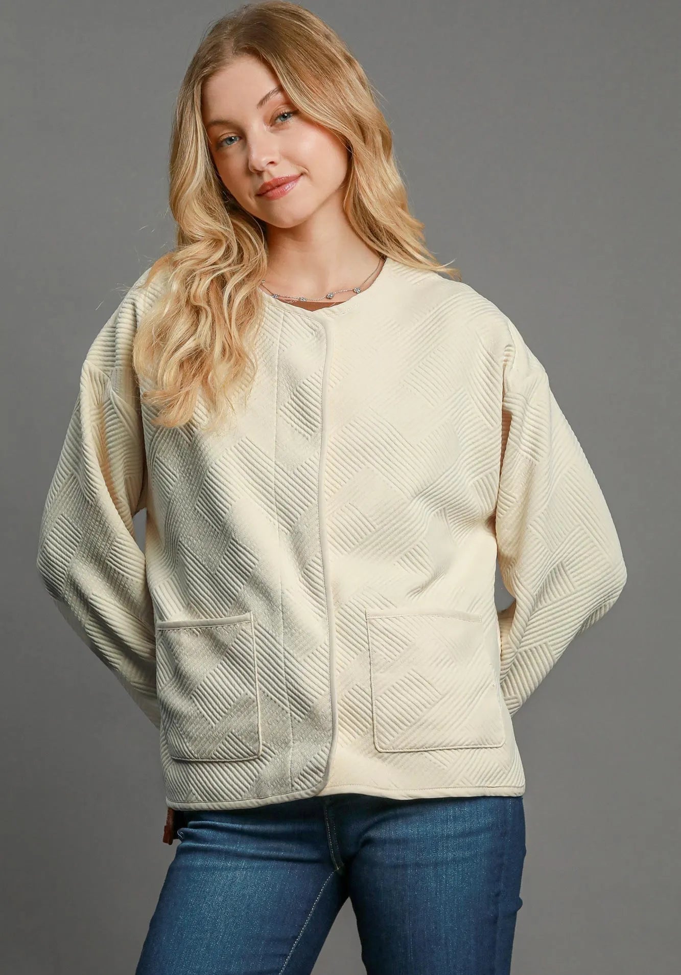 Woman wearing a cream-colored textured jacket with blue jeans on a gray background