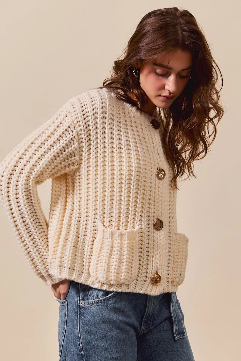 Woman wearing a chunky knit beige sweater with gold buttons on a beige background