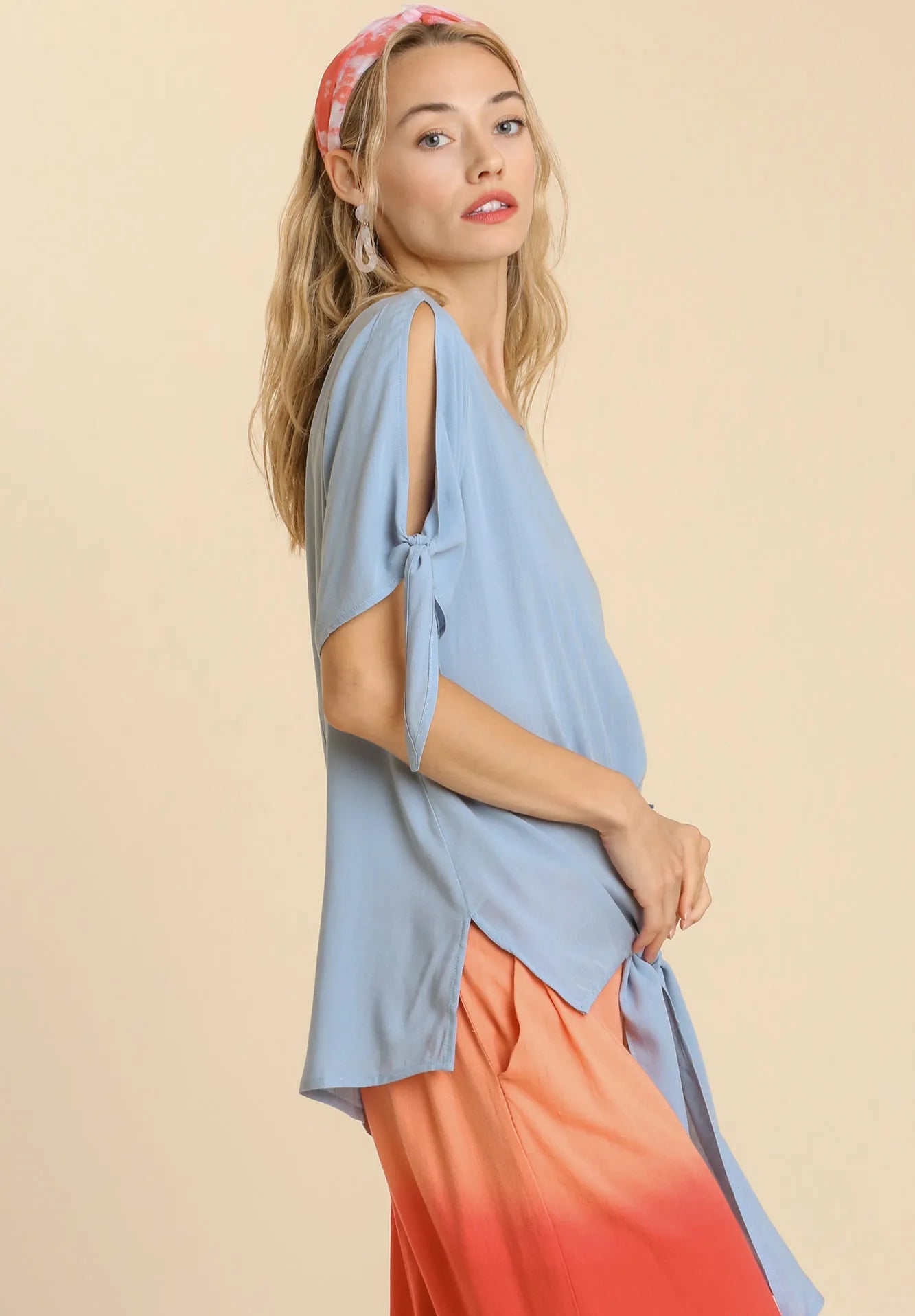 Woman wearing a dusty blue top with a bow detail and orange pants on a beige background