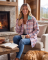 Beach Rose Co. pink and blue plaid shacket with a cozy sherpa collar worn by a woman sitting by a fireplace.