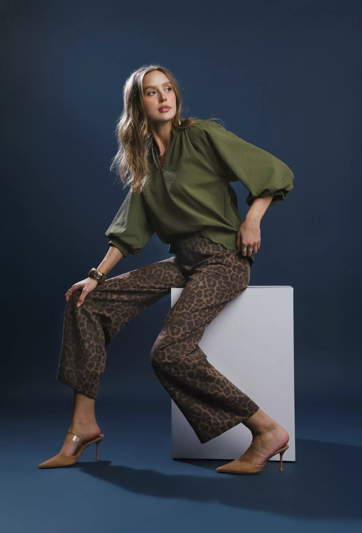 Woman in green top and leopard print pants sitting on a white block against a dark blue background