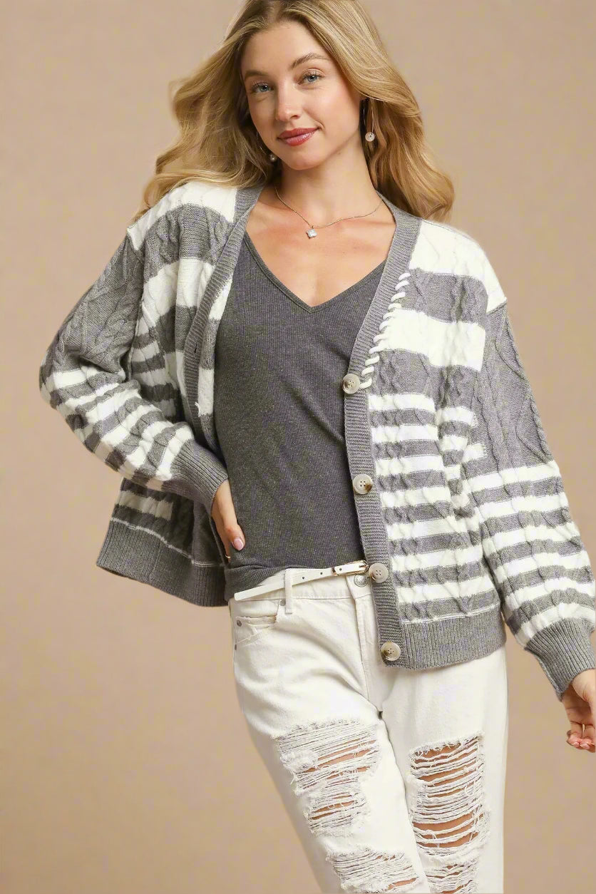 Woman wearing a gray and white striped cardigan over a gray shirt and white pants on a beige background.