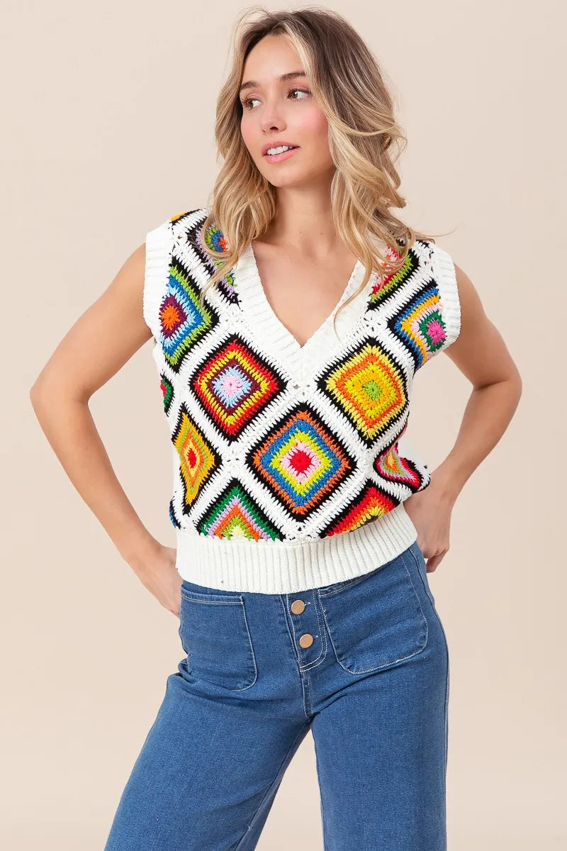 Woman wearing a colorful crochet vest over a white shirt with blue jeans on a beige background