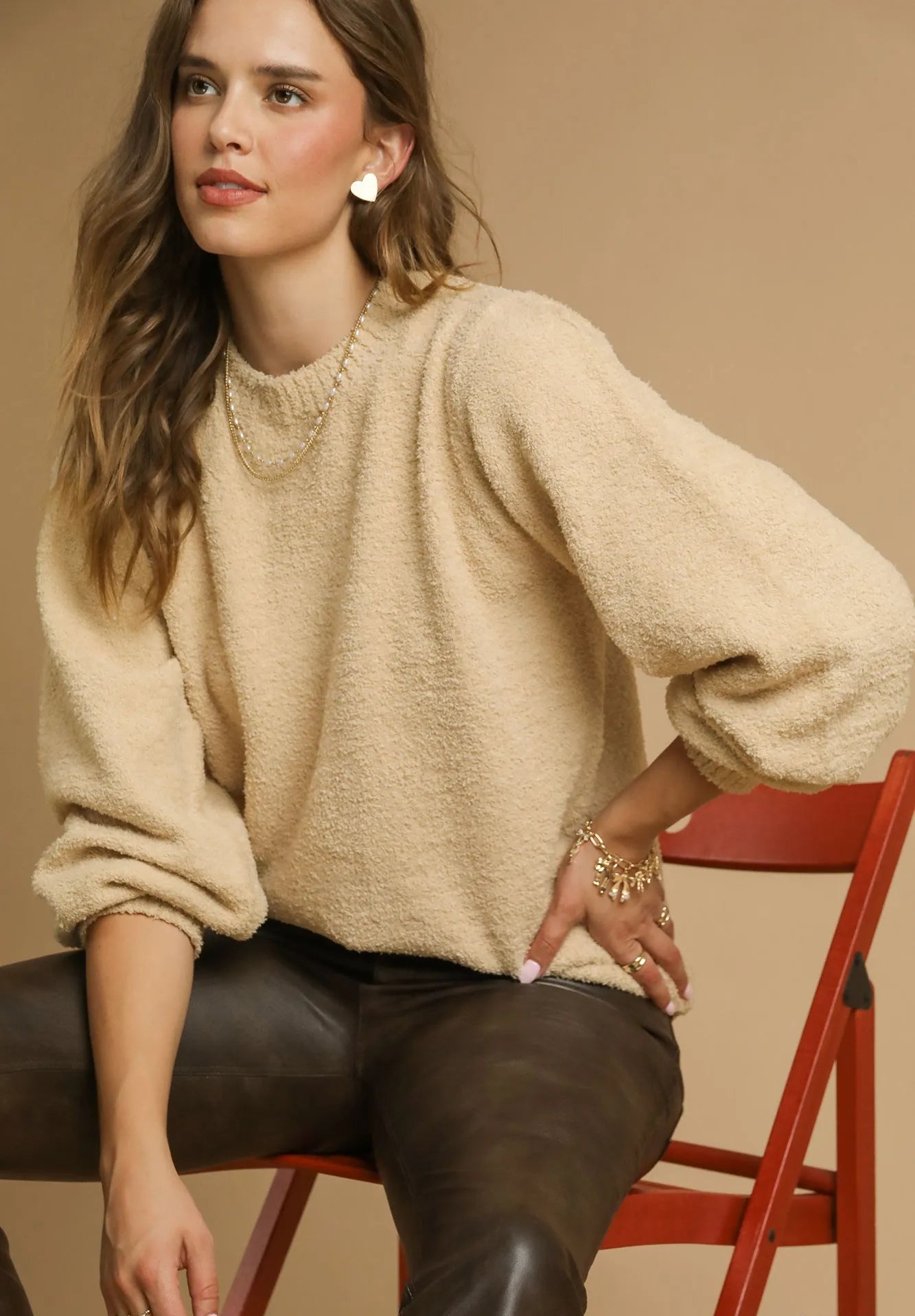 Woman wearing a beige sweater and brown pants sitting on a red chair against a beige background.