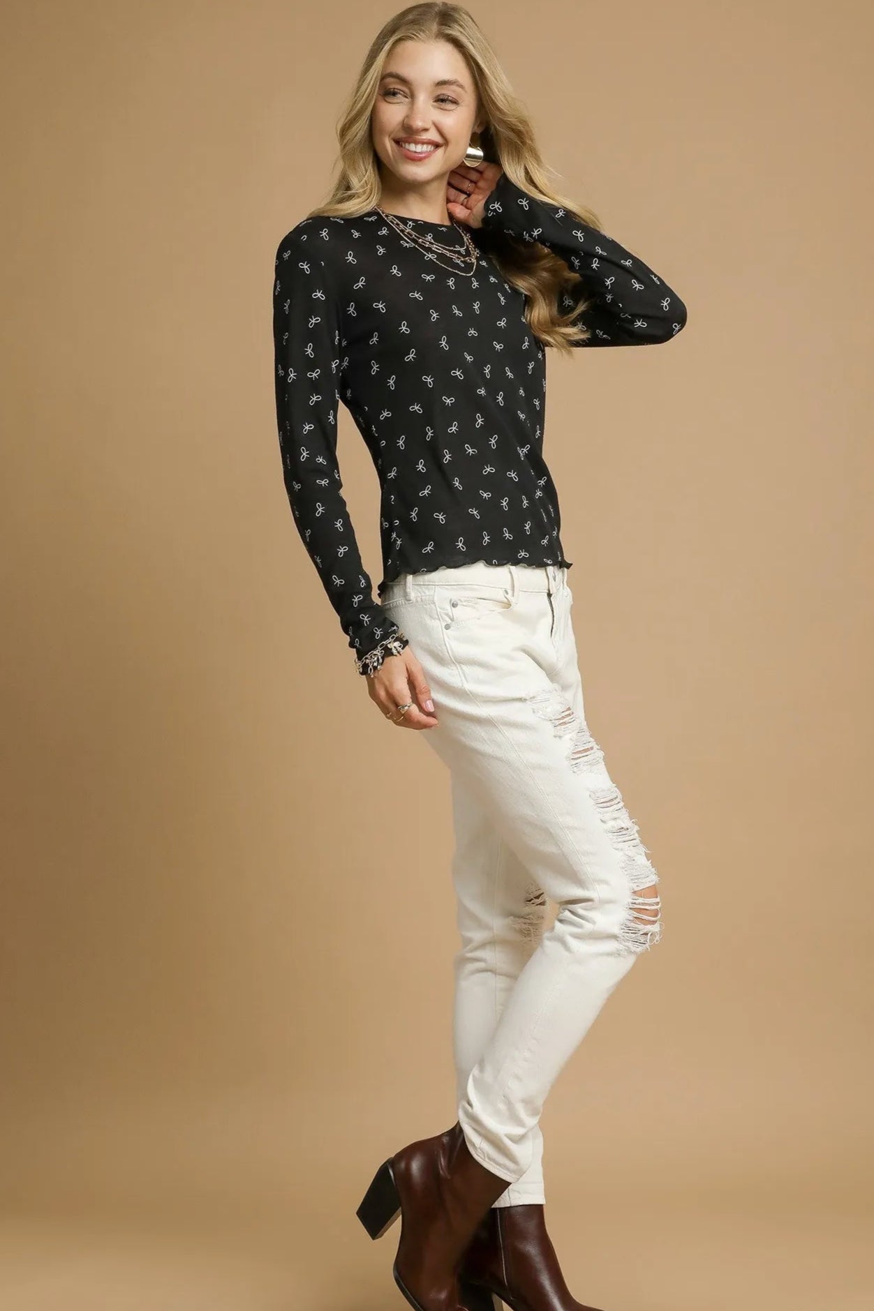 Woman wearing a black patterned top, white pants, and brown boots on a beige background