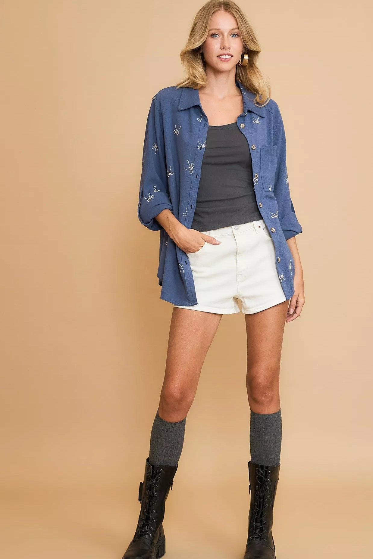 Woman wearing a denim shirt, black top, white shorts, and black boots on a beige background