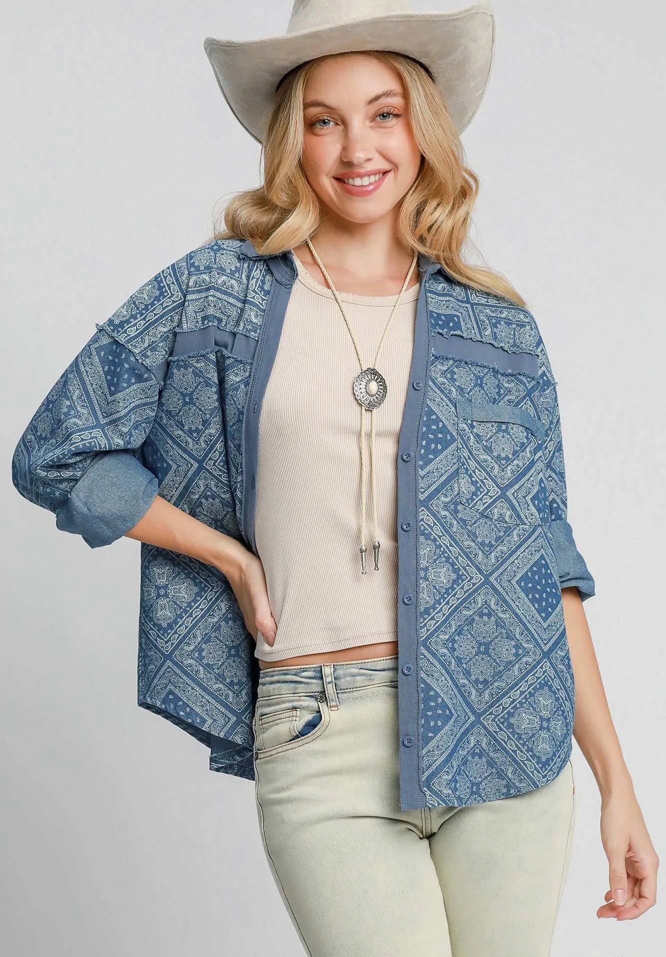 Woman wearing a blue patterned denim jacket, beige top, light green jeans, and a beige cowboy hat on a white background.