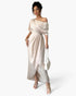 A woman shown from the back wearing a floor-length off-shoulder gown in a light warm beige or cream tone against a plain light gray studio background.