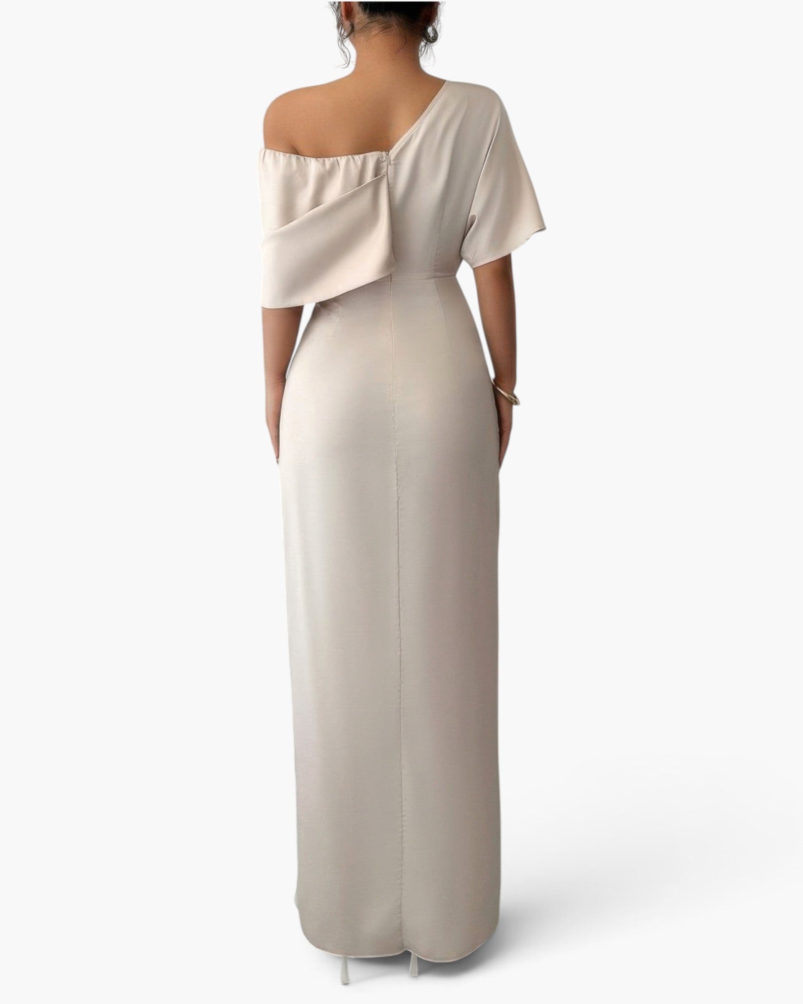 A woman shown from the back wearing a floor-length off-shoulder gown in a light warm beige or cream tone against a plain light gray studio background.
