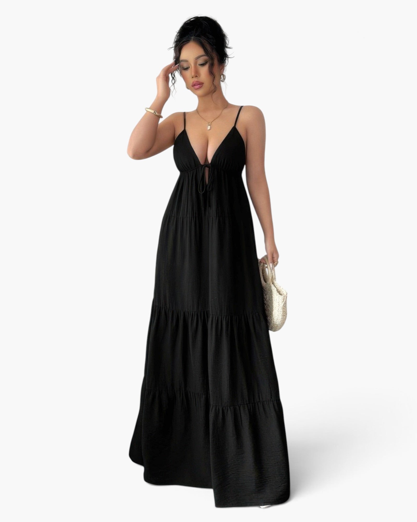 A woman modeling a black spaghetti-strap maxi dress with a tiered skirt and a keyhole tie-front detail, holding a small white handbag against a light gray studio background.