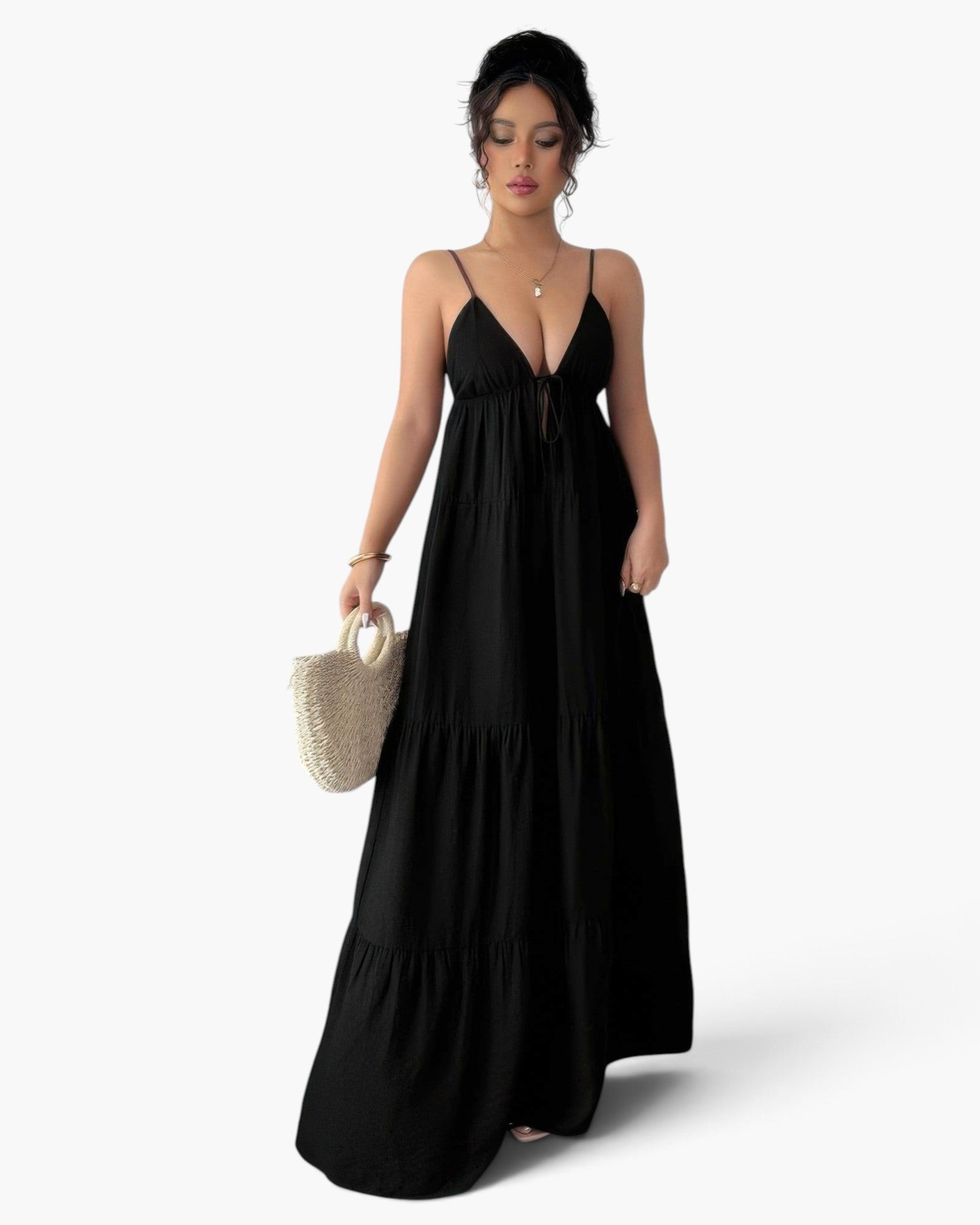 A woman modeling a long black spaghetti-strap maxi dress with a tiered skirt, holding a small woven straw handbag against a plain white studio background.