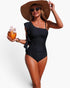 Beach Rose Co. black one-piece swimsuit with asymmetrical ruffle shoulder, worn by a woman with a sun hat and coconut.