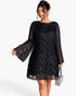 A woman modeling a black sheer lace dress with long bell sleeves, fringe trim, and a textured chevron pattern, holding a small silver handbag against a plain white background.