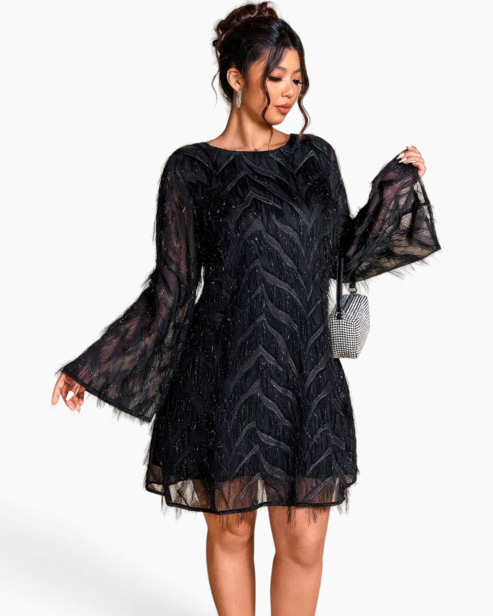 A woman modeling a black sheer lace mini dress with long bell sleeves, sequined chevron patterns, and a flared silhouette, paired with a small silver handbag against a plain white background.