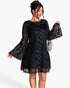 A woman modeling a black sheer lace mini dress with long bell sleeves, sequined chevron patterns, and a flared silhouette, paired with a small silver handbag against a plain white background.