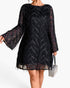 A woman wearing a black sheer lace dress with long bell sleeves and a chevron pattern, holding a small rhinestone handbag against a plain white background.