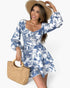 A woman modeling a blue and white toile-print romper with long puff sleeves, a sweetheart neckline, and a ruffled hem. She is wearing a wide-brimmed straw hat and holding a woven straw handbag against a clean white background.